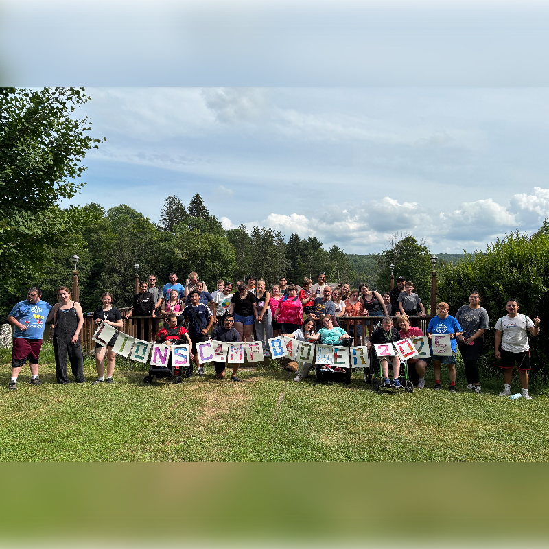 A large group of people, some in wheelchairs, pose for a photo holding a banner that reads "LIONS CAMP BUDDY 2022" in front of a lush green landscape with trees and a cloudy sky.