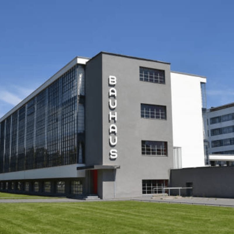 A modern Bauhaus building with large glass windows and the word "BAUHAUS" in large letters stands on a manicured green lawn, with other buildings visible in the background under a clear blue sky.