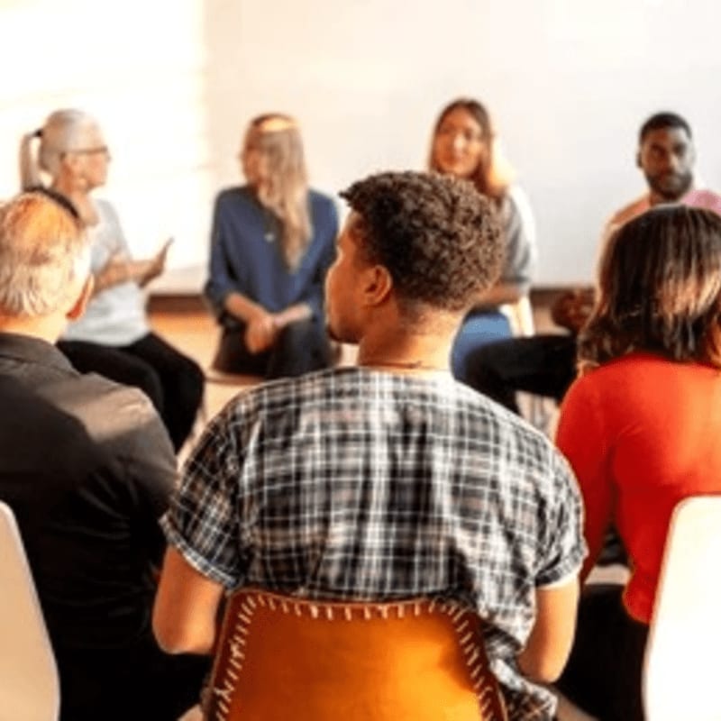 In the foreground, a young man with curly hair in a plaid shirt sits facing away from the camera, while in the background, a diverse group of people are seated in a circle, engaged in conversation.