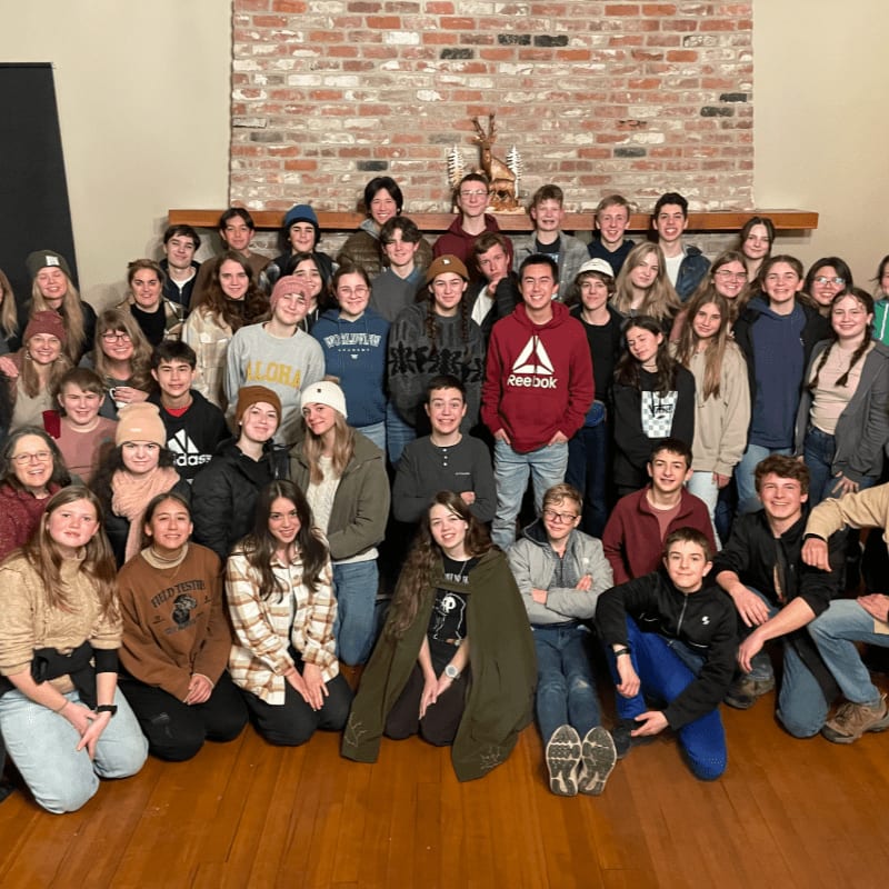 A large group of young people are gathered in front of a brick fireplace, with some sitting on the wooden floor and others standing behind them.