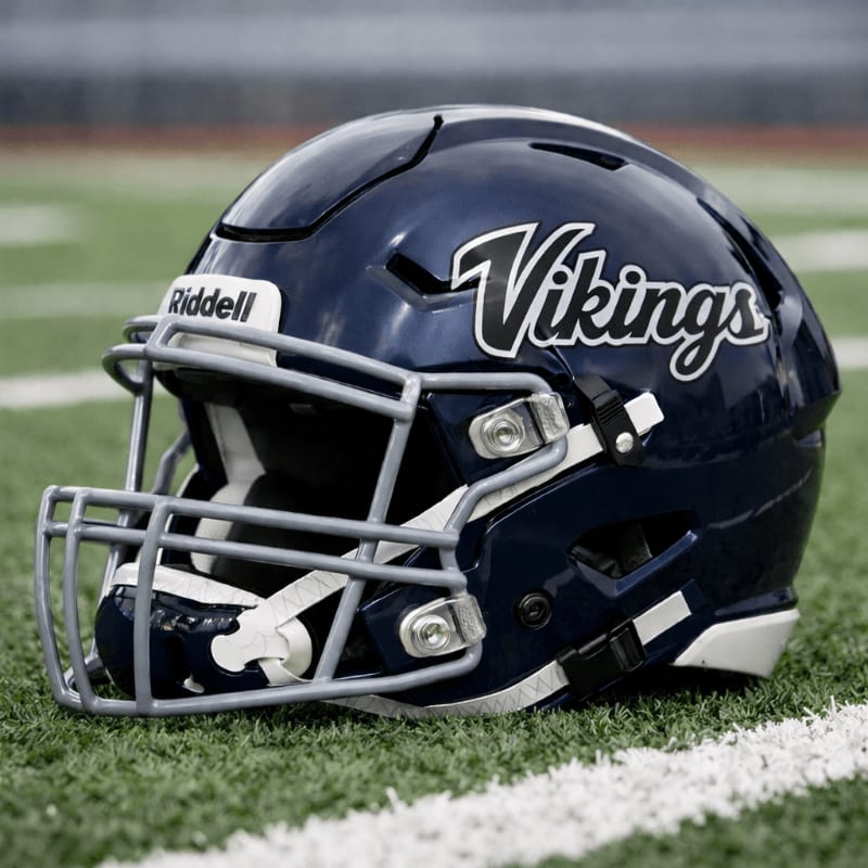 A dark blue "Vikings" football helmet rests on a green field with white yard lines, while a blurred stadium stands in the background.