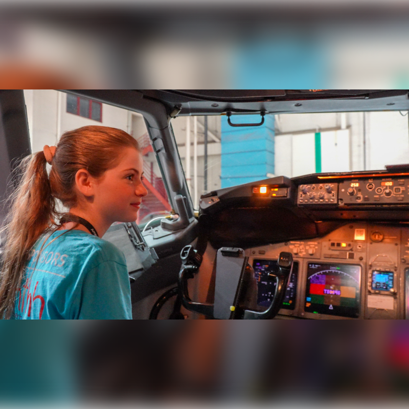 A young girl with a ponytail in a blue shirt sits in the cockpit of a plane, looking at the illuminated control panel.