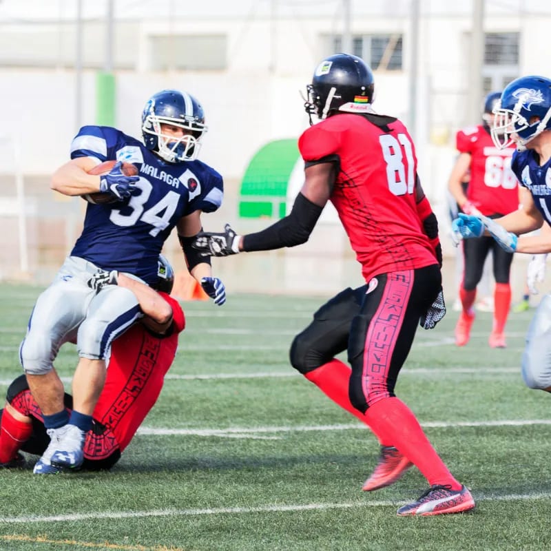 In the foreground, an American football player in a blue jersey with the number 34 is tackled by an opponent in a red jersey, while in the background, other players in similar uniforms are visible on a grassy field under a bright sky.