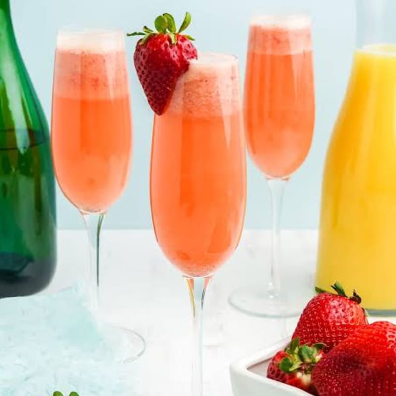 Three glasses of strawberry mimosa with a strawberry garnish are in the foreground, with a green bottle and a carton of orange juice in the background.