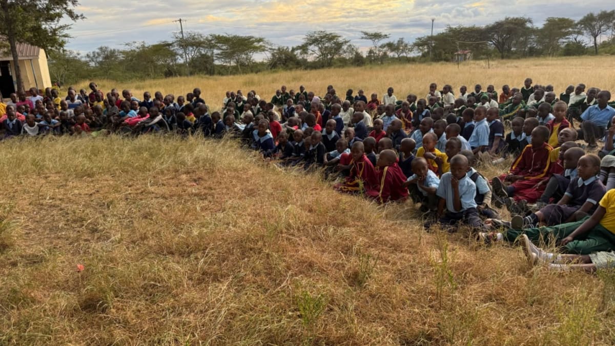 A large group of children sit in rows on dry grass in the foreground, facing a building and trees in the hazy background.