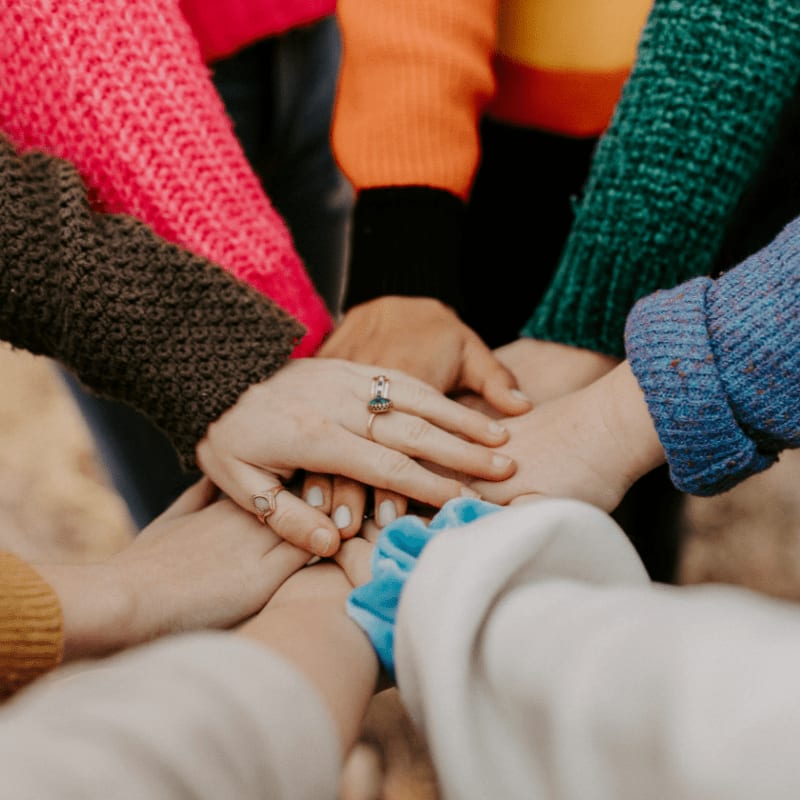 A pile of hands in colorful sweaters are stacked in the foreground, with a blurred background.