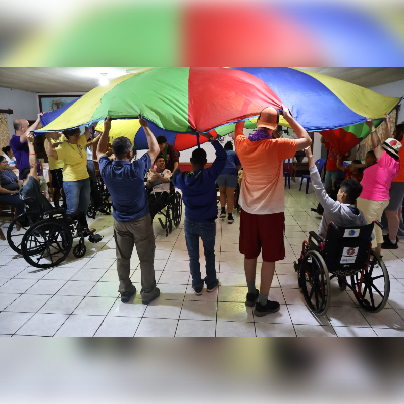 People in wheelchairs and standing hold up a large, colorful parachute in a room with tiled floors and a light-colored ceiling.