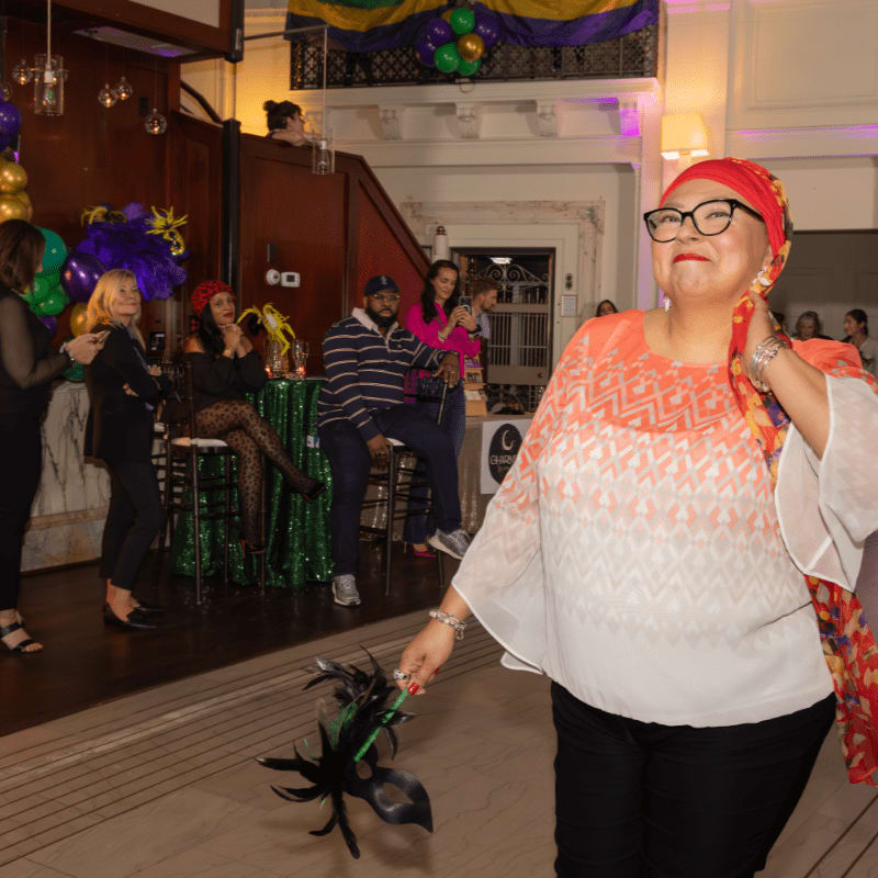 A woman in the foreground holds a mask, while in the background, people mingle at a festive event with Mardi Gras decorations.