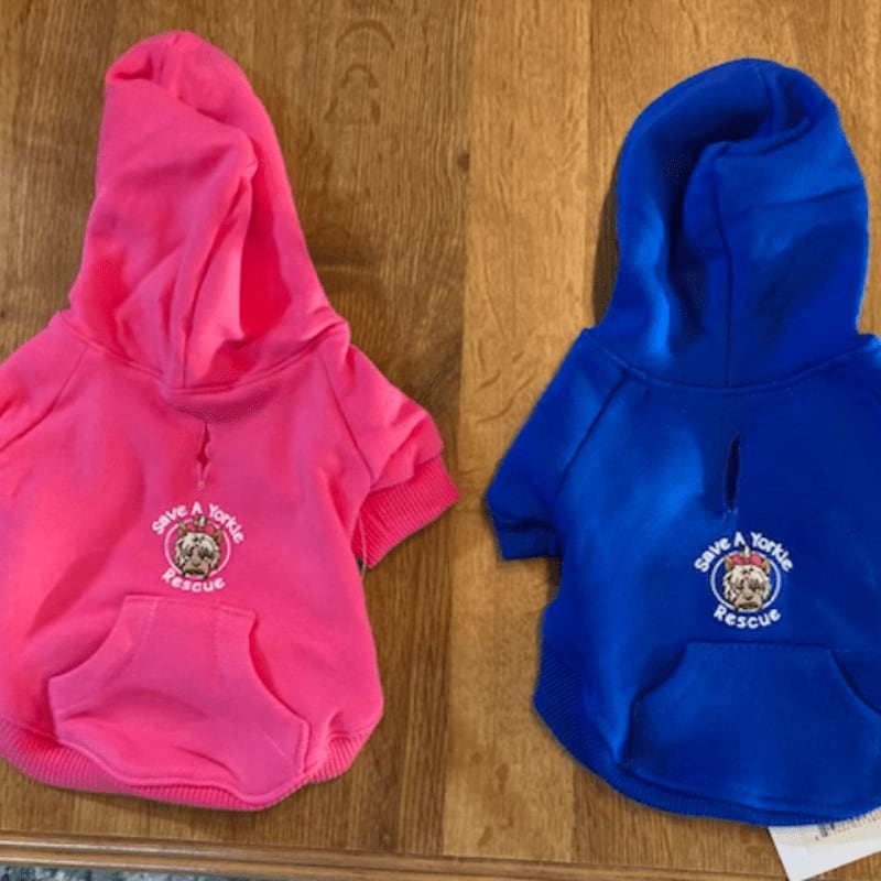 Two dog hoodies, one pink and one blue, with "Save a Yorkie Rescue" logos are laid out on a wooden surface.