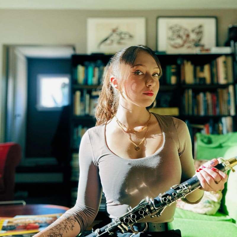 A young woman with a clarinet in her hands sits in front of a bookshelf filled with books.