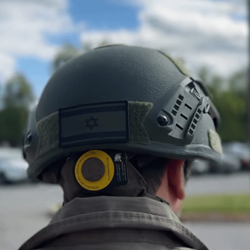 A close-up of a dark green tactical helmet with an Israeli flag patch is worn by a person, with a blurred background of trees and parked cars under a cloudy sky.