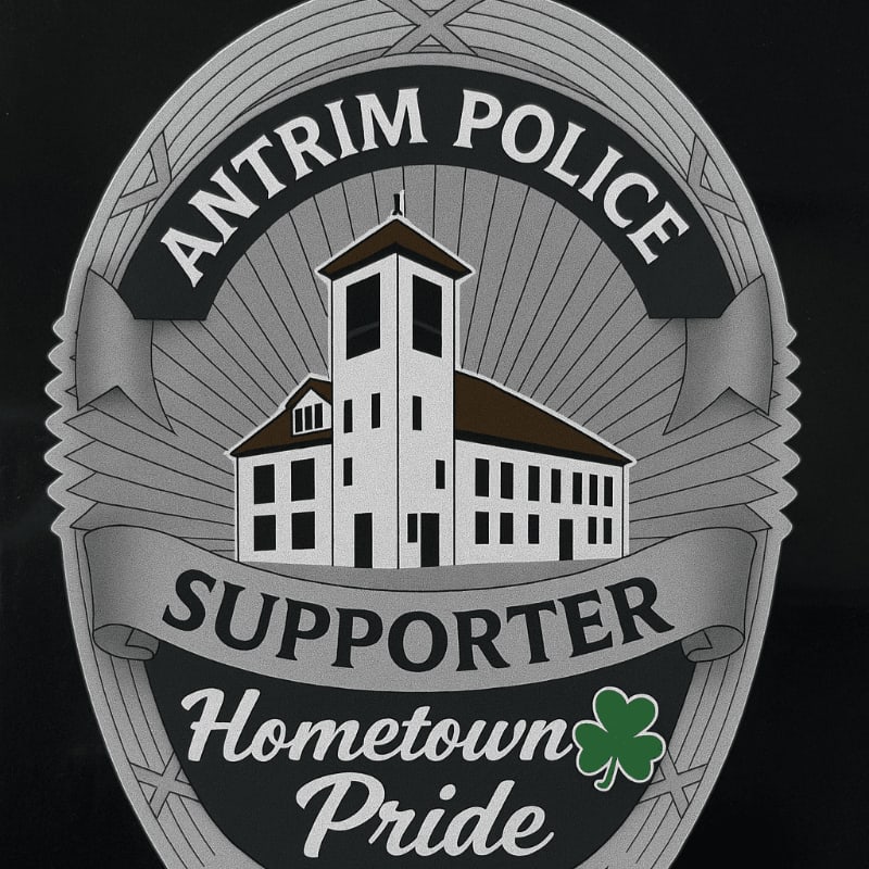 A silver badge with a white building and the words "Antrim Police Supporter Hometown Pride" is set against a black background.