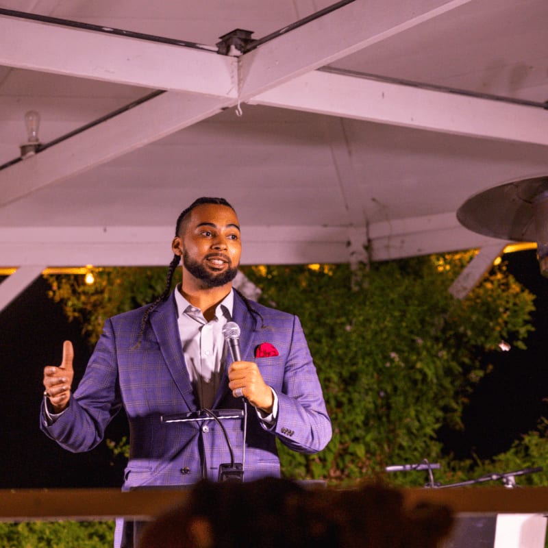 A man in a suit speaks into a microphone in the foreground, while string lights and greenery are visible in the background.