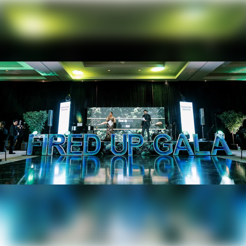 Two people stand on a stage in front of large, illuminated "FIRED UP GALA" lettering, with a blurred, reflective floor in the foreground and a dark, draped background with spotlights.