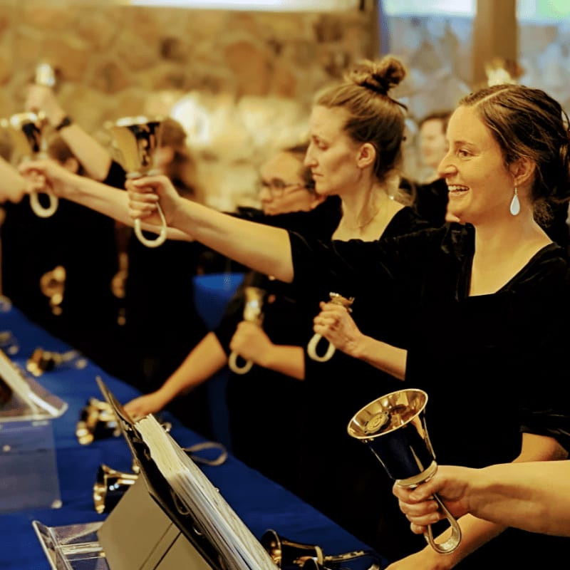 A group of women in black dresses ring handbells in front of a stone wall.