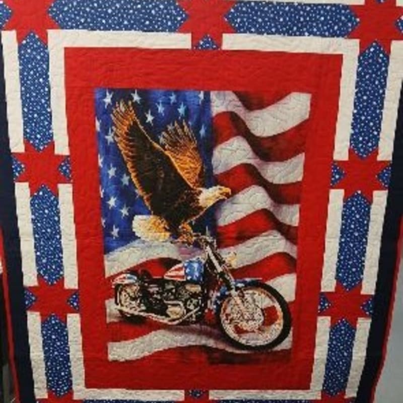 A bald eagle soars over a motorcycle in front of a waving American flag, all framed by a patriotic quilt border.