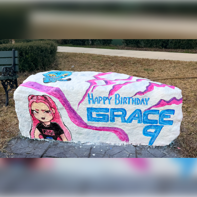 A large white rock painted with a cartoon character and "Happy Birthday Grace 9" sits on a paved area in front of a park bench and manicured bushes.