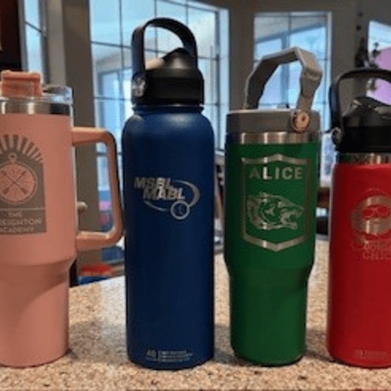Four colorful insulated water bottles, two pink and blue on the left and two green and red on the right, stand on a speckled countertop with a blurred background of a room.