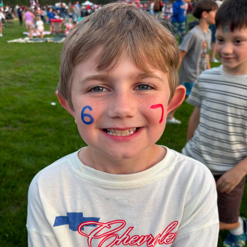 A young boy with face paint of the numbers 6 and 7 smiles at the camera in the foreground, while a blurred crowd enjoys an outdoor event in the background.