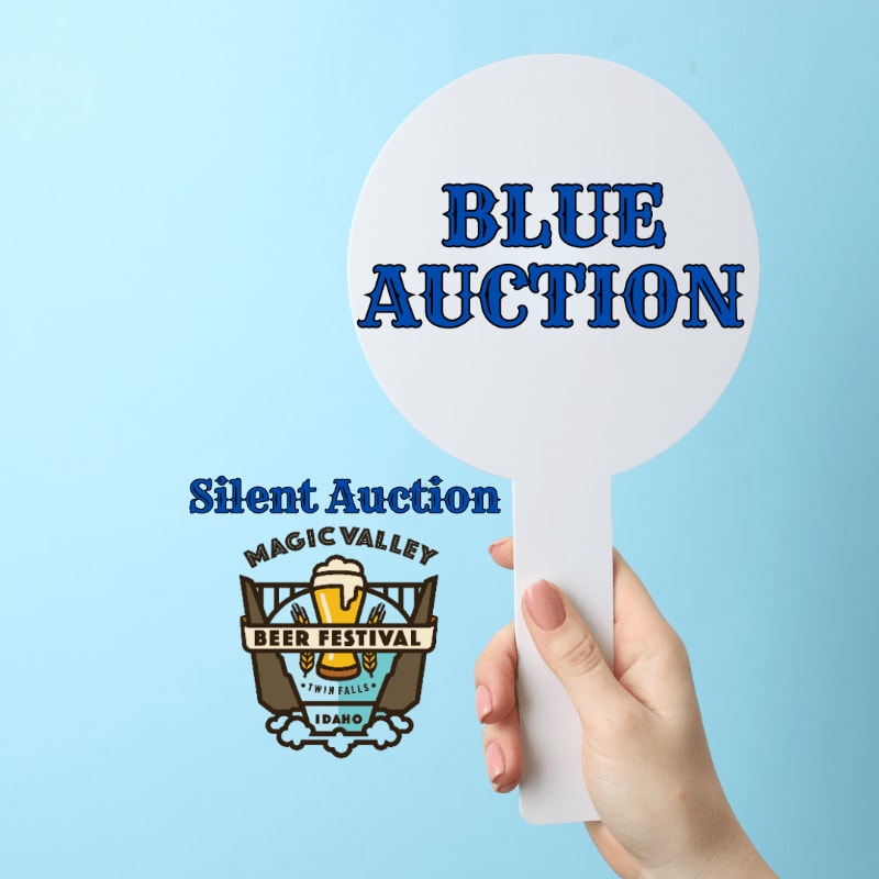 A hand holds a white paddle with "BLUE AUCTION" written in blue text against a light blue background, with a "Silent Auction Magic Valley Beer Festival" logo to the left.