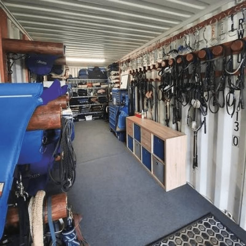 Blue saddle pads and horse tack hang on wooden racks in the foreground, while shelves filled with equestrian gear line the corrugated metal walls of the storage container in the background.