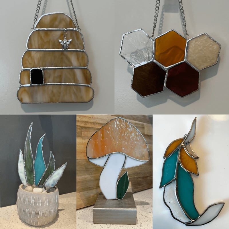 Stained glass art pieces, including a beehive, honeycomb, succulent, mushroom, and feather, hang against a plain wall and sit on a countertop.