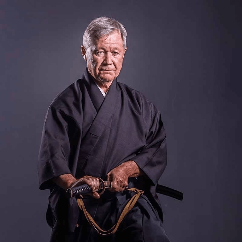 An elderly man in a dark gi holds a katana in front of a plain gray background.
