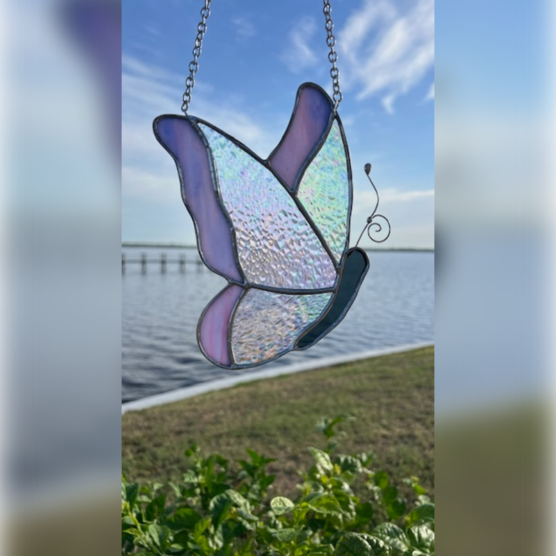 A stained glass butterfly hangs in front of a serene blue sky and calm water, with lush green foliage in the foreground.