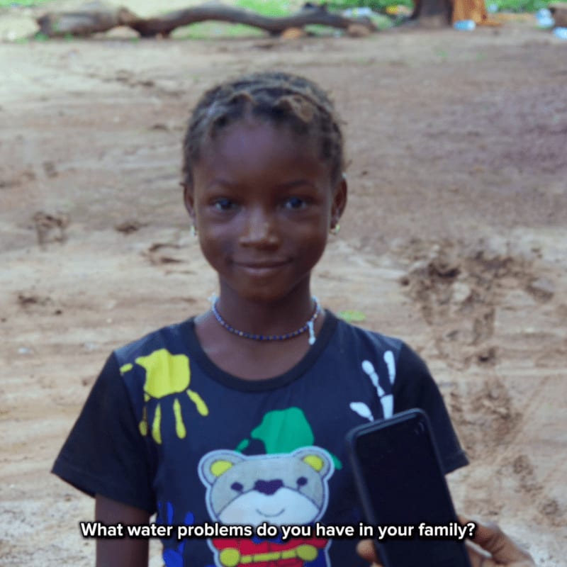 A young girl with braided hair smiles at the camera in the foreground, while a blurred background shows a dirt path and trees.