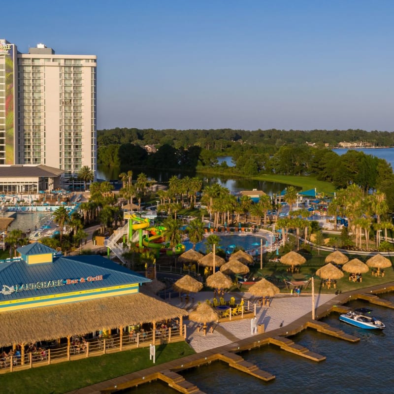 A waterfront resort with a thatched-roof restaurant in the foreground and a tall hotel and lush trees bordering a lake in the background.