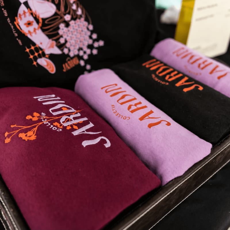 A burgundy t-shirt with an orange floral design is in the foreground, with three other folded t-shirts in shades of lavender, black, and purple behind it, all featuring the "JARDIN COLLECTIF" logo.