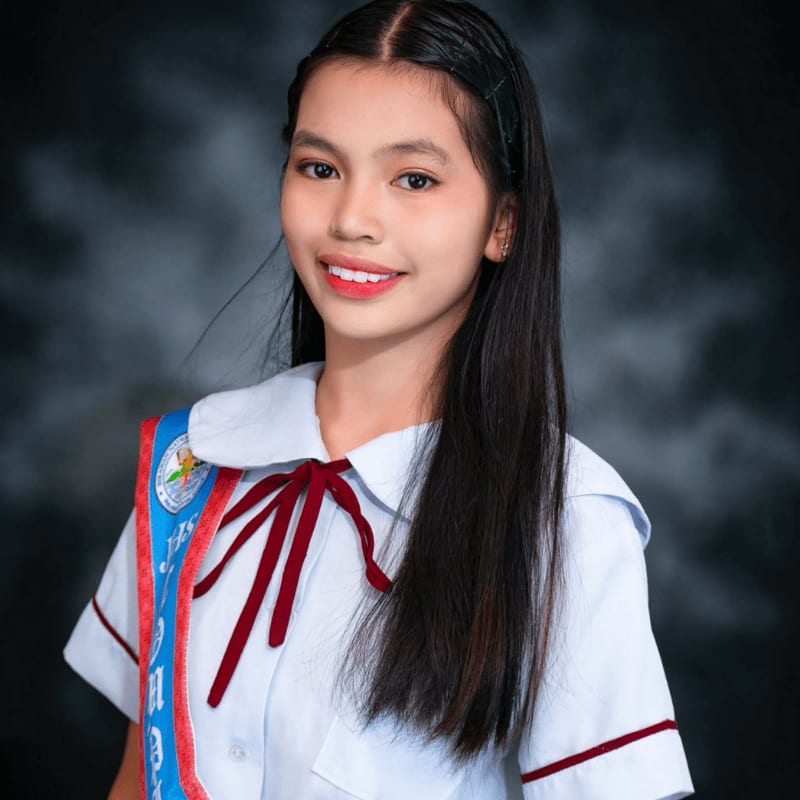 A young woman in a school uniform with a sash smiles against a dark, textured background.