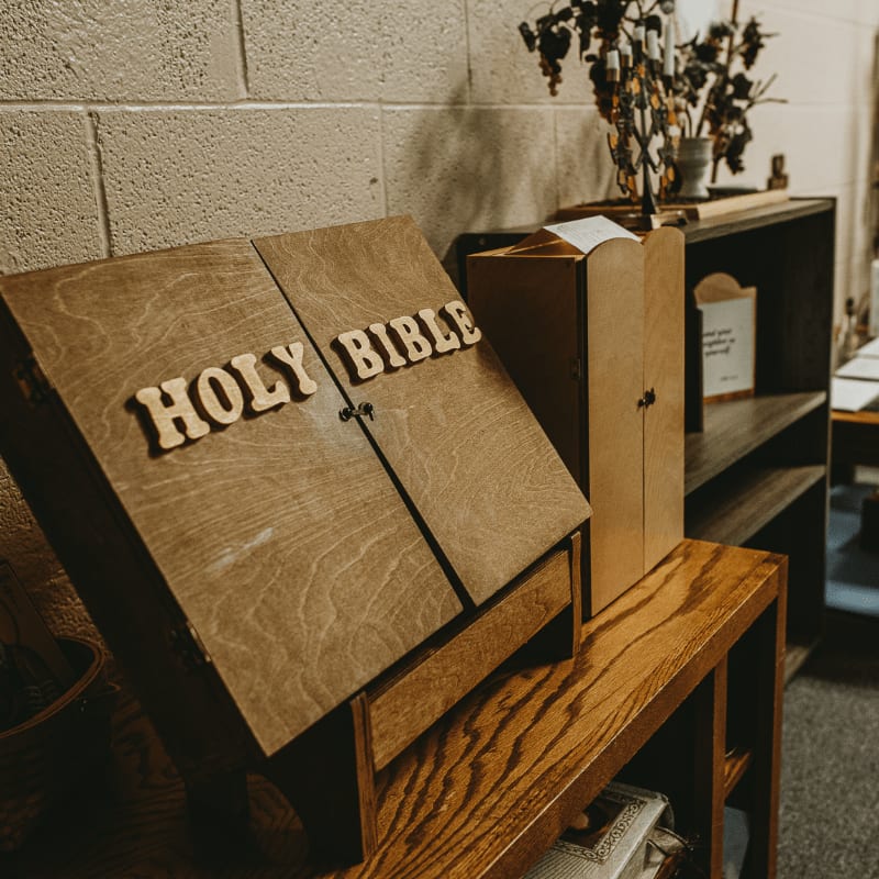 A wooden Bible stand with "HOLY BIBLE" carved into it sits on a table in the foreground, with shelves and decorative items in the background.
