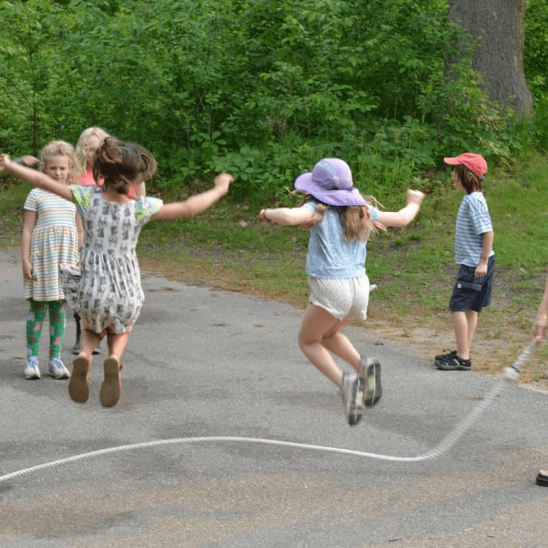 Children in the foreground jump rope on a paved path, while a lush green forest forms the background.