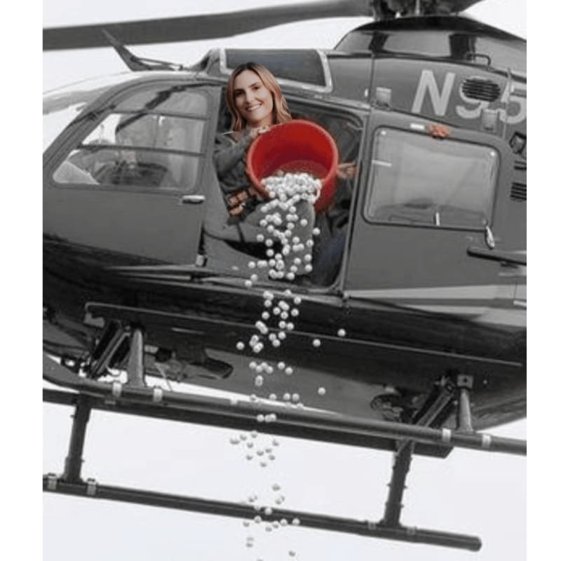 A woman in a helicopter in the foreground pours white balls from a red bucket, with a black and white helicopter in the background.