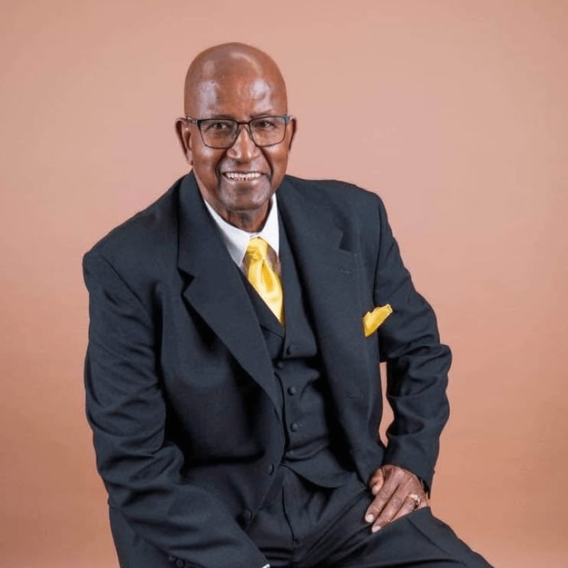 A smiling, elderly Black man in a suit and tie sits against a plain, light brown background.