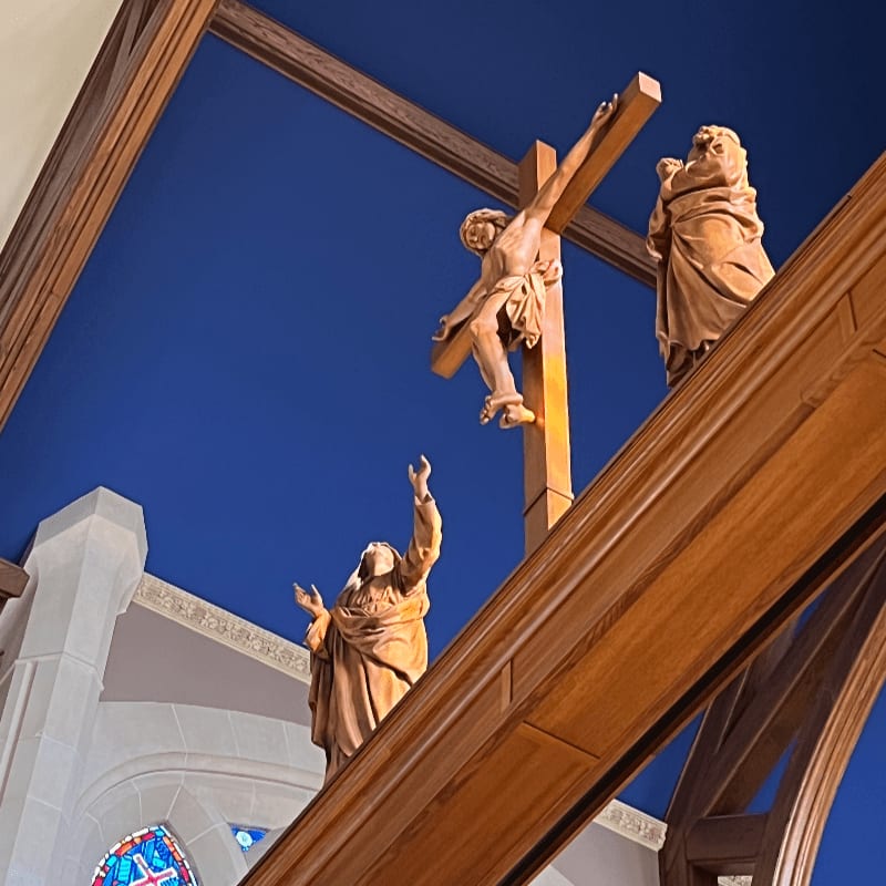 Wooden statues of Jesus on the cross and two other figures are set against a deep blue background, with architectural elements of a church visible in the lower left.