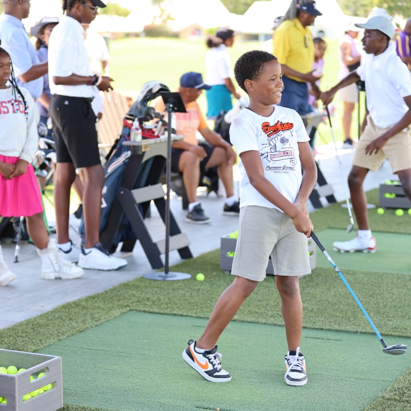 A young boy in the foreground swings a golf club on a practice green, while other children and adults watch and practice in the blurred background.