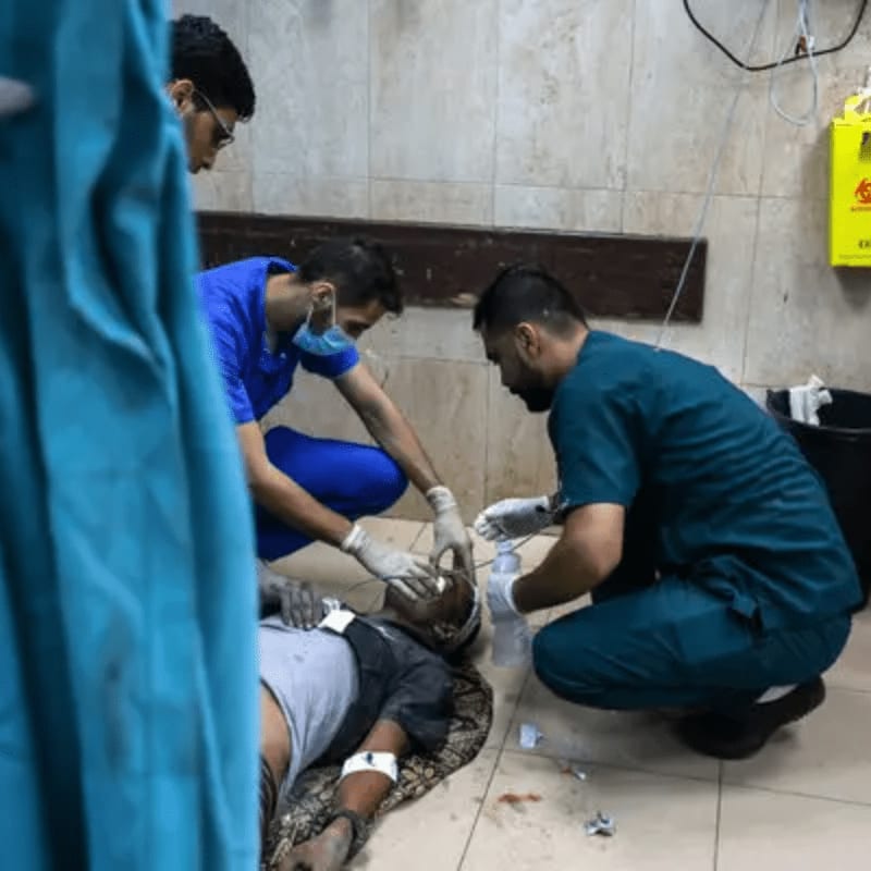 Two medical professionals in scrubs attend to an injured person lying on the floor, with a blue curtain in the foreground and a sterile room with medical equipment in the background.