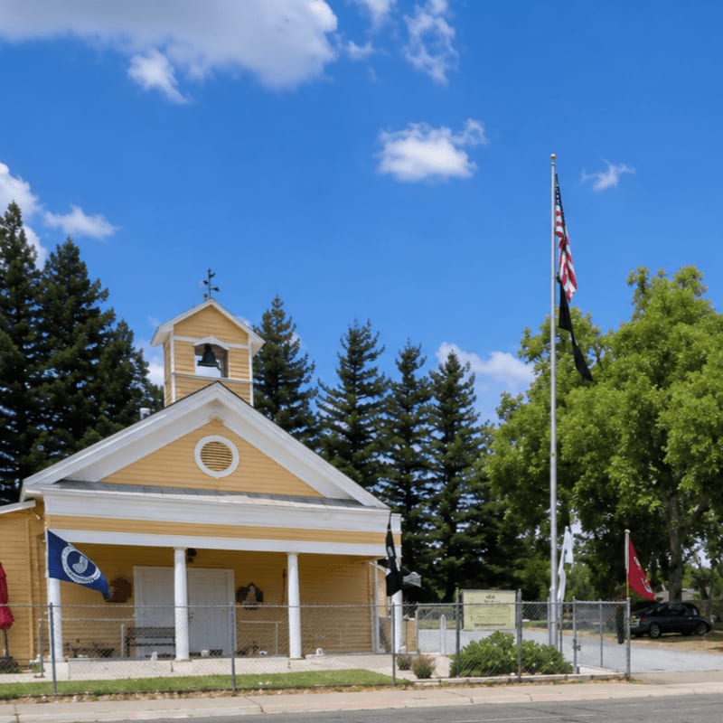 A yellow building with a bell tower stands behind a chain-link fence adorned with flags, set against a backdrop of green trees and a bright blue sky with scattered clouds.