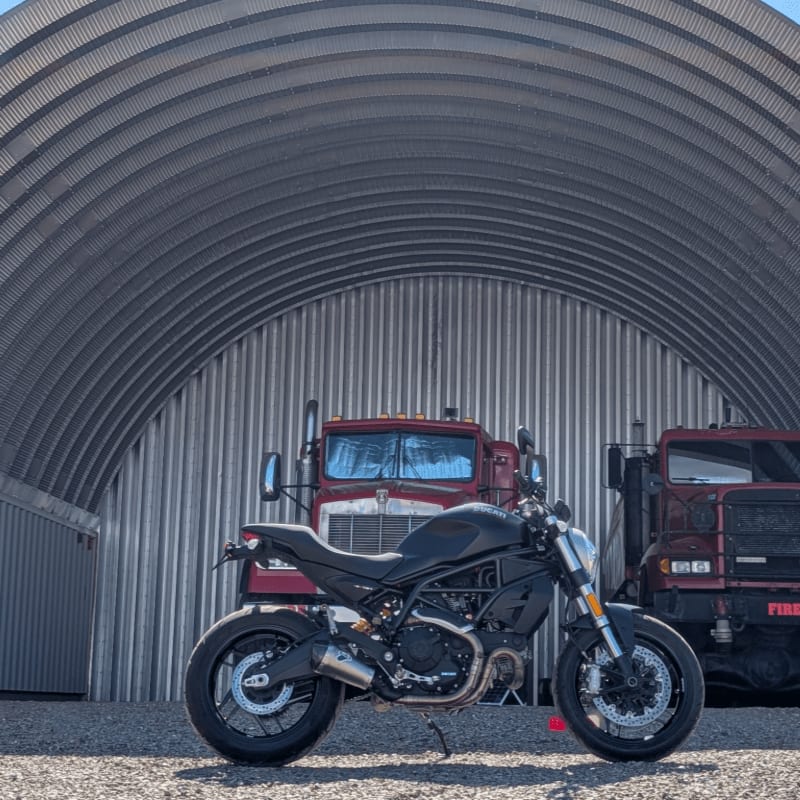 A black motorcycle is parked in front of two red trucks inside a large, corrugated metal Quonset hut.