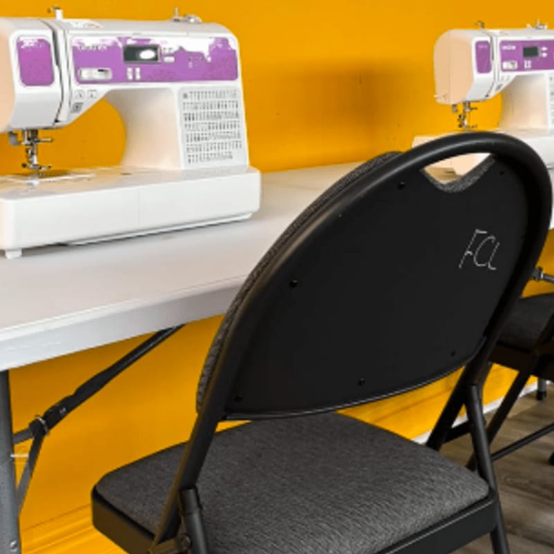A black folding chair with "FCL" written on its back is in the foreground, with two white sewing machines on a table in the background against a bright yellow wall.
