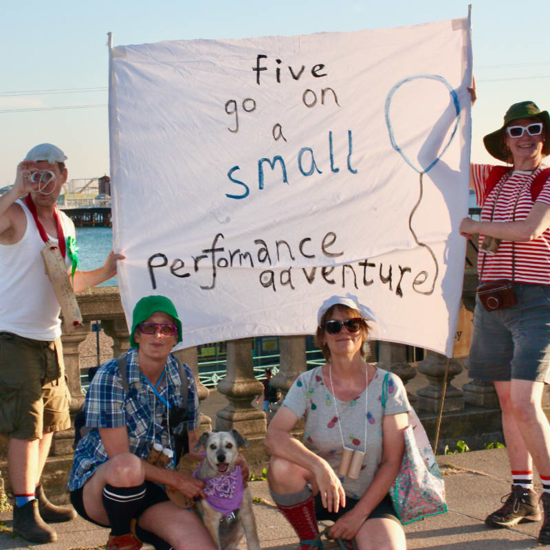 Four people and two dogs pose in front of a banner that reads "five go on a small performance adventure," with a blurred cityscape in the background.