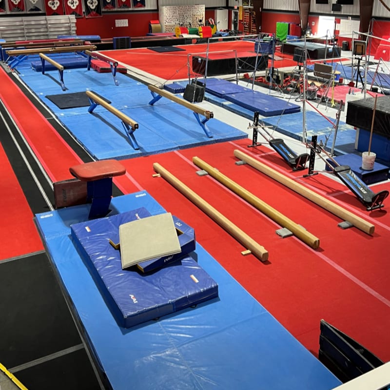 In the foreground, blue mats and balance beams are laid out on a red floor, while the background reveals a spacious gymnastics gym with various equipment.