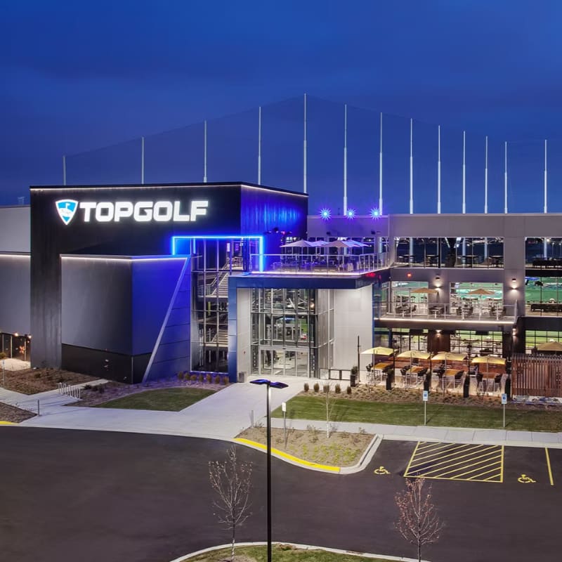 A modern Topgolf entertainment complex with a parking lot in the foreground and illuminated netting stretching into the twilight sky in the background.