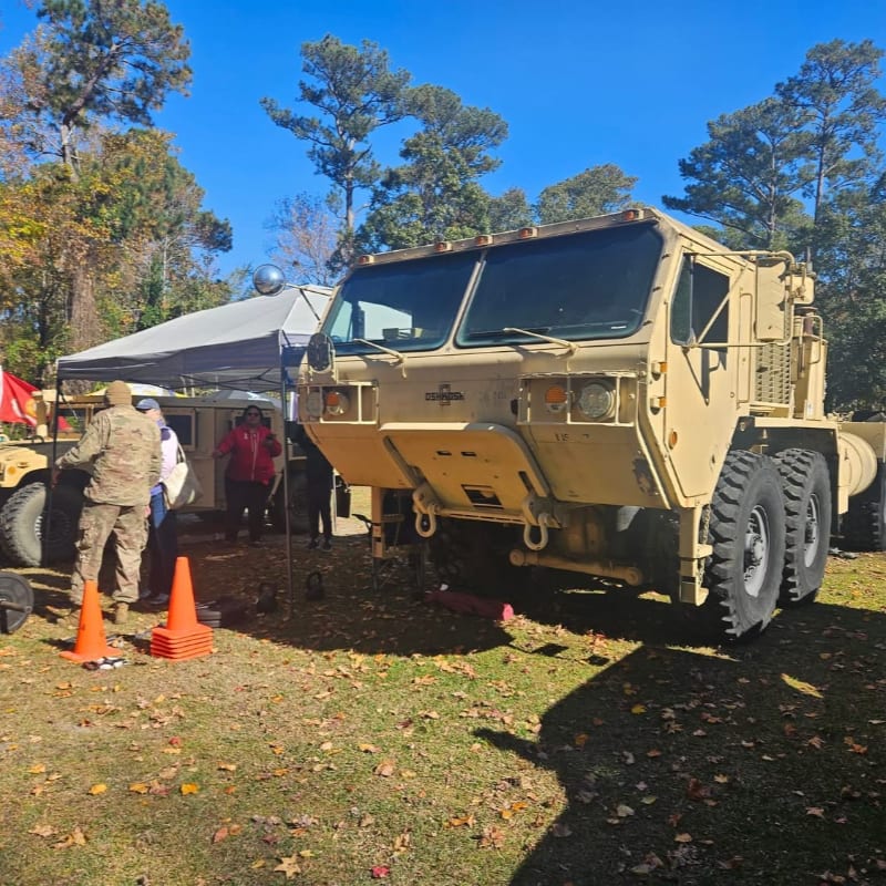 A tan military truck is parked on a grassy field with people and a tent in the background.