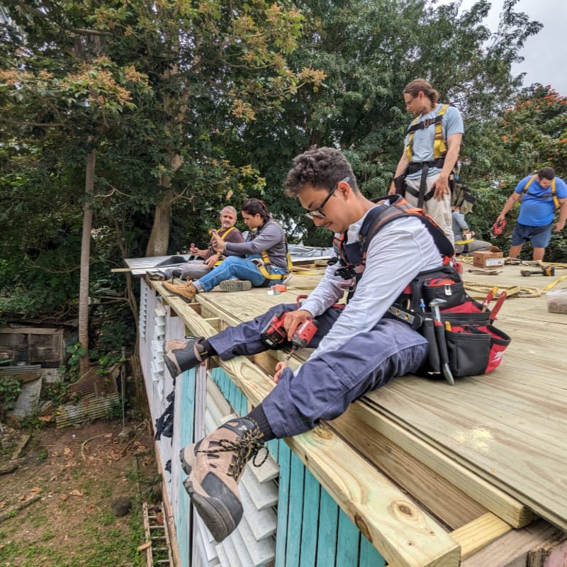 A man in the foreground uses a drill on a wooden roof while others work in the background amidst lush green trees.