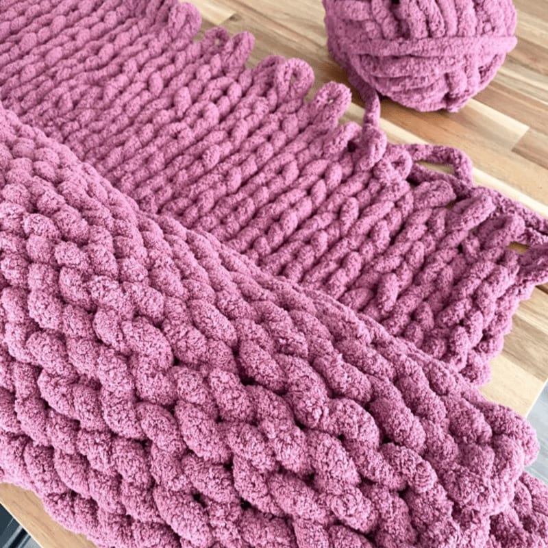 A chunky knit scarf in a dusty rose color lies on a wooden surface, with a matching ball of yarn in the background.