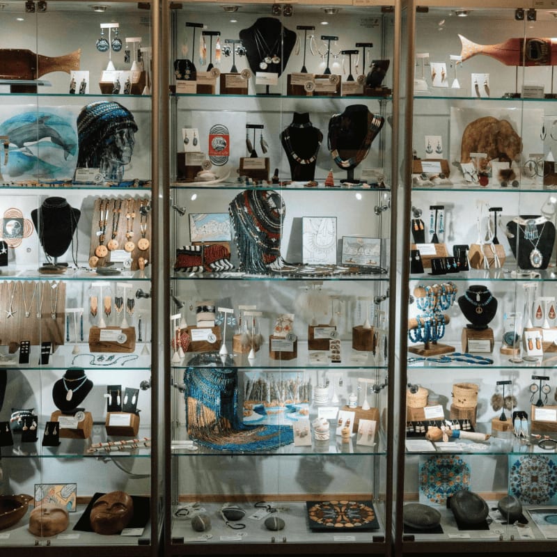 In the foreground, a variety of handcrafted jewelry and art pieces are displayed on shelves, while the background features more items behind glass in a retail setting.