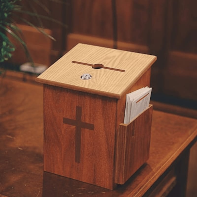 A wooden offering box with a cross carved into its side sits on a polished wooden table, with blurred pews and greenery in the background.